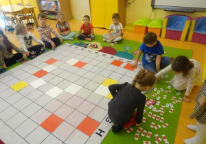 Troje dzieci wybiera z pośród rozłożonych na dywanie kodów zapisanych na karteczkach. Na środku dywanu leży mata do kodowania wypełniona kilkoma kolorowymi płytkami, Reszta dzieci siedzi wokół maty i przygląda się.